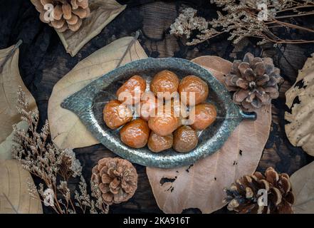 Drei aromatisierte, konservierte chinesische Pflaumen (konservierte Früchte) auf einem kleinen, silberblattförmigen Teller. Sein Geschmack ist sauer, salzig und süß. Draufsicht, SE Stockfoto