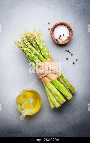 Frisches Spargelbündel, Zutaten zum Kochen: Salz, Pfeffer, Zitrone und Olivenöl auf grauem Küchentisch-Hintergrund, Draufsicht, Kopierraum Stockfoto