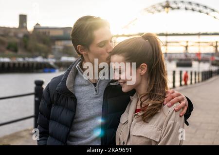 Ein junges Paar, das zusammen den Tag in Newcastle upon Tyne verbringt. Sie gehen auf einem gepflasterten Gehweg entlang des Flusses Tyne und umarmen sich gegenseitig Stockfoto