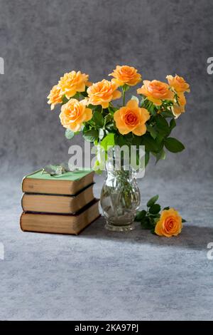 Strauß cremefarbener Rosen in einer Kristallvase, drei Bücher und Gläser auf grauem Hintergrund Stockfoto