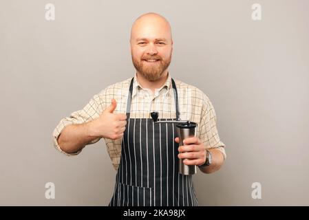 Ein junger kahler Barista hält eine Kaffeemühle fest und empfiehlt sie mit einem Daumen nach oben auf hellgrauem Hintergrund. Stockfoto