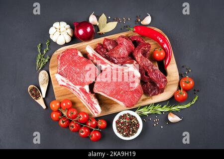 Auswahl an rotem Fleisch - rohes Ribeye-Rindfleisch-Steaks und gehacktes rohes Rind- oder Lammfleisch, bereit zum Grill mit Gewürzen und Gemüse auf Holzschneidschweinen Stockfoto