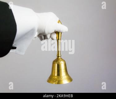 Isoliertes Bild der weißen Hand mit Goldglocke auf weißem Hintergrund. Ring für Servicekonzept. Professionelle Gastfreundschaft und Eleganz. Stockfoto