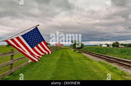 An einem wolkigen Tag winken amerikanische Flaggen im Wind, auf einem Zaun in der Nähe einer Single Railroad Track Stockfoto