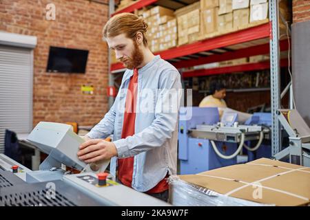 Waist up Porträt von bärtigen jungen Mann Einrichtung Druckmaschine in der Werkstatt, Kopierer Raum Stockfoto