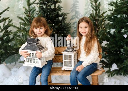 Zwei lustige kleine Schwestern halten Laternen und sitzen auf einer Bank zwischen den Weihnachtsbäumen Stockfoto