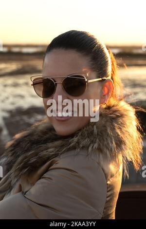 Porträt einer jungen kaukasischen Frau in goldenem Mantel und Sonnenbrille, die lächelnd auf die Kamera blickt. Lifestyle-Konzept Stockfoto