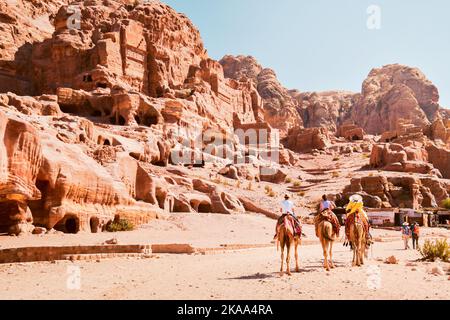 Lokaler arabischer Reiseführer in der Stadt Petra, der auf Kamelen reitet, mit Touristen, die an sonnigen, heißen Tagen an königlichen Petra-Gräbern vorbei fahren Stockfoto