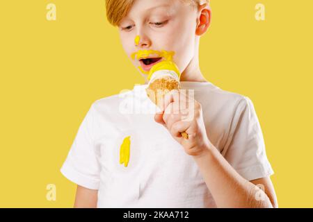 Kind verschüttet Eis. Die Kleidung ruiniert. Ein Junge, der Bananeneis auf gelbem Hintergrund isst. Isoliert Stockfoto