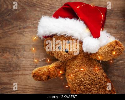 Teddybär im Weihnachtsmannhut. Plüschtier mit Glühbirnen auf schäbigen Holzhintergrund. Neujahr. Stockfoto