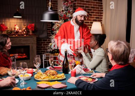 afroamerikanische Frau bekommt Geschenk von Mann in weihnachtsmann Kostüm, Familie Austausch weihnachtsgeschenke. Freunde feiern Heiligabend, essen traditionelle Mahlzeit am festlichen Tisch Stockfoto