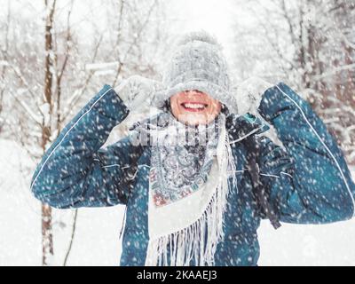 Lächelnde Frau spielt mit gestricktem Hut. Spaß im verschneiten Winterwald. Die Frau lacht, als sie durch den Wald geht. Aufrichtige Emotionen. Stockfoto