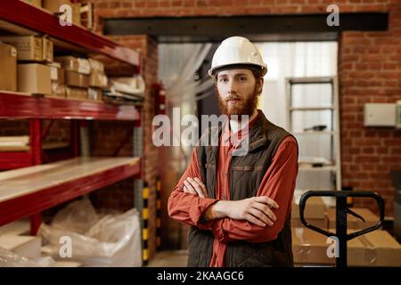 Porträt eines bärtigen Arbeiters, der im Fabriklager einen Hardhut trägt und mit gekreuzten Armen auf die Kamera blickt Stockfoto