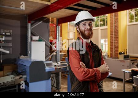 Porträt eines bärtigen Arbeiters, der in einer Fabrik mit Hartschlüpfeln unterwegs ist und mit gekreuzten Armen auf die Kamera blickt Stockfoto