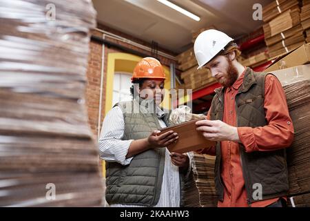 Porträt von zwei Arbeitern, die in der Werkshalle Hardhuts tragen und ein digitales Tablet verwenden Stockfoto