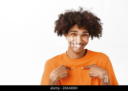 Nahaufnahme von niedlichem, gen-z Kerl mit lockigen Haaren und Piercing, zeigt auf sich selbst und lächelt, lachend, stehend über weißem Hintergrund Stockfoto
