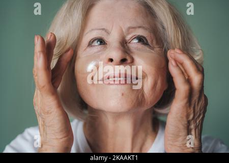 Nahaufnahme einer pensionierten Frau, die kosmetische Eingriffe mit Augenflecken unter isolierten Augen über der grünen Wand vorführt. Pflege der trockenen Haut. Schönheitstherapie Stockfoto