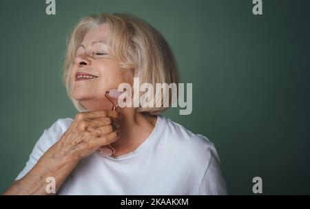 Beauty Face Portrait einer hübschen älteren Frau, die ihr Gesicht mit einer Jadewalze massiert, ihre Augen schließt, isoliert über einer grünen Wand. Natürliche Anti-Falten Stockfoto