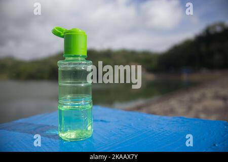 Eukalyptusöle in einer transparenten grünen Flasche auf blauem Beton mit See- und tropischen Bergen-Hintergrund, in kreativem Schießstil. Stockfoto