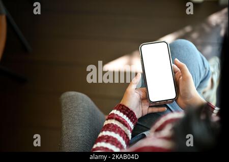 Oben sehen, schöne junge asiatische Frau mit ihrem Smartphone, während entspannt sitzt auf einem Sessel im Wohnzimmer. Zugeschnittenes Bild Stockfoto