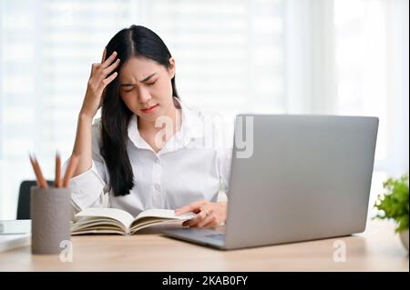 Müde und gestresste junge asiatische Geschäftsfrau, die an Kopfschmerzen oder Migräne leidet, während sie im Büro an ihrem Projekt arbeitet. Stockfoto