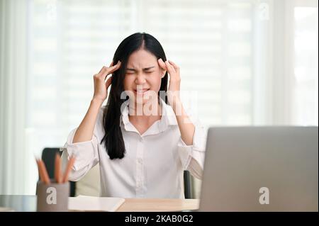 Gestresste und wütende junge asiatische Geschäftsfrau, die am Schreibtisch im Büro sitzt, Kopfschmerzen hat, mit ihrem Projekt unzufrieden ist und einen großen Profi hat Stockfoto
