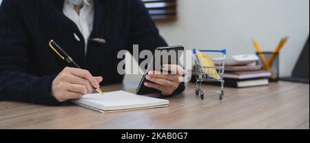 Frau schreiben in Notebook Verwenden Sie Handy Studie online zu Hause. Glückliche ethnische weibliche Notizen handschriftlich auf Papier mit Anwendung auf dem Smartphone. Planung Stockfoto