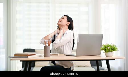 Überarbeitete junge asiatische Geschäftsfrau oder Büroangestellte, die an Nackenschmerzen litt, nachdem sie einen langen Tag bei der Arbeit hatte und ihren Hals massierte Stockfoto