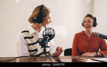 Lächelnde Podcast-Moderatorin interviewt einen Gast in ihrem Heimstudio. Zwei glückliche Frauen, die eine gute Zeit haben, während sie eine digitale Audioübertragung für ihre s aufnehmen Stockfoto