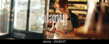 Glückliche Besitzerin eines Supermarkts, die ein digitales Tablet benutzt, während sie in ihrem Supermarkt steht. Erfolgreiche Unternehmerin, die ihr kleines Unternehmen mit Wireless betreibt Stockfoto