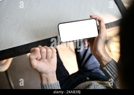 Über dem Kopf geschossen, ein asiatischer Mann hält ein Smartphone weißen Bildschirm Mockup in horizontaler Position und zeigt geballte Faust. Stockfoto