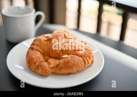 Köstliches frisches französisches Croissant mit Mandelscheibe auf einem weißen Teller, serviert mit heißem Kaffee oder Kakao. Café-Bar oder Gebäck der Bäckerei Stockfoto