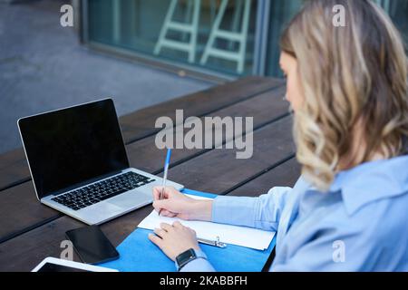 Nahaufnahme eines Porträts einer Studentin, die an Online-Kursen teilnimmt, mit einem Laptop an der frischen Luft sitzt und sich Notizen macht. Videochat für Geschäftsfrau Stockfoto