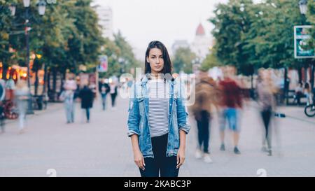 Porträt einer müden jungen Studentin, die allein im Stadtzentrum steht und mit geradem Gesicht die Kamera anschaut, während sich Männer und Frauen in der Stadt herumschwirrend bewegen. Stockfoto