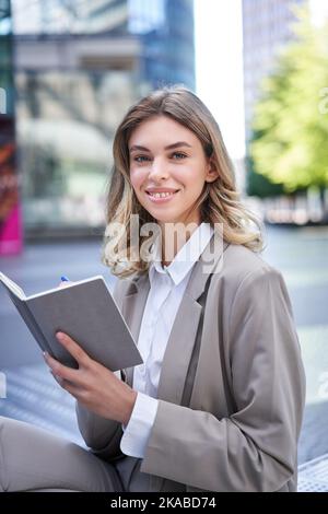 Eine junge Frau im Firmenanzug sitzt mit Notizbuch und Stift, notiert sich, arbeitet und schreibt ihre Ideen auf Stockfoto