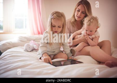 Eine App am Tag hält die Langeweile fern. Ein kleines Mädchen nutzt ein digitales Tablet, während es sich mit ihrer Mutter und Schwester auf dem Bett verklebt. Stockfoto