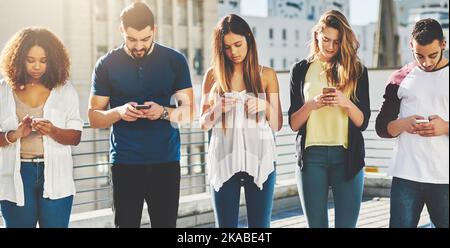 Wir leben in einer technologiebasierten Gesellschaft. Eine Gruppe junger Menschen, die im Freien auf ihrem Handy SMS schreiben. Stockfoto