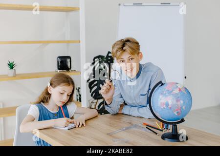 Nette Schüler, die in der Schule den Globus im Unterricht betrachten. Junge hält Lupe und schaut zur Kamera Stockfoto
