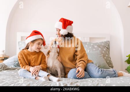 Frohe Weihnachten und Frohes neues Jahr. Glückliche Familie mit Hund Jack russell warten auf das neue Jahr in santa claus Hüte, sitzen Seite an Seite auf dem Bett zu Hause im Schlafzimmer. Mama und glückliche Tochter in Pullover Stockfoto