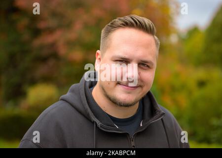Freundlicher, lächelnder junger Mann mit Schnurrbart und Bart, der vor einem Garten posiert Stockfoto