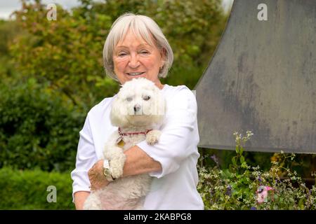 Ältere grauhaarige Dame hält ihren süßen kleinen weißen Hund liebevoll in den Armen Stockfoto