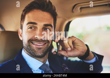 Er machte seinen Erfolg allein: Ein fröhlicher junger Geschäftsmann, der sich auf seinem Handy unterhielt, während er auf dem Rücken eines Autos saß. Stockfoto