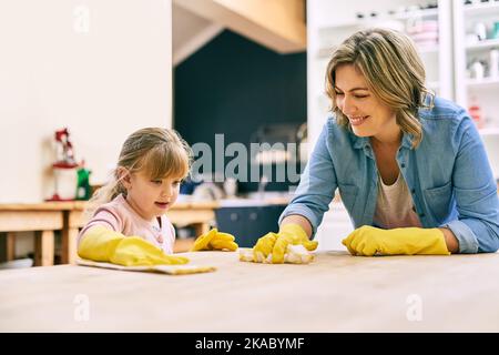 Du hast den Dreh raus, mein kleines Mädchen. Eine fröhliche junge Mutter und ihre kleine Tochter putzen gemeinsam einen Tisch. Stockfoto