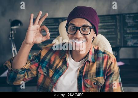 Portrait von fröhlich positive Hacker Kerl sitzen Stuhl Handfinger demonstrieren Okey Symbol Workstation drinnen Stockfoto
