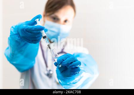 Junge Ärztin in einer medizinischen Maske und Handschuhen mit einer Spritze in der Hand und einem Stethoskop in einem Krankenhaus während einer Coronavirus-Pandemie. Selektive Stockfoto