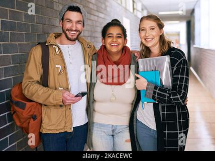 Freunde vervollständigen das Universitätserlebnis. Beschnittenes Porträt von drei jungen Universitätsstudenten, die in einem Korridor auf dem Campus stehen. Stockfoto