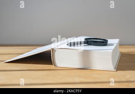 Lupe mit offenem dickem Buch auf Holzschreibtisch mit Tageslicht. Stockfoto