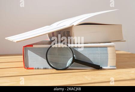 Lupe in der Nähe eines Stapels dichter und offener Bücher auf einem Holzschreibtisch mit Tageslicht. Stockfoto