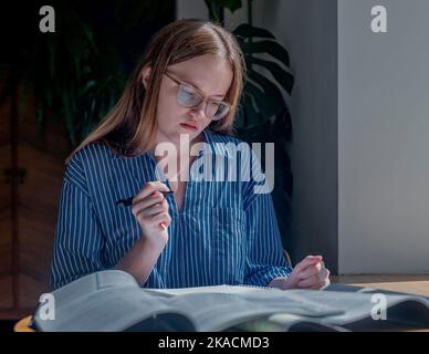 Junge Frau in der Brille denkt, liest, bereitet sich mit Notizbüchern auf Prüfungen vor, Bücher am Holzschreibtisch im Café. Stockfoto