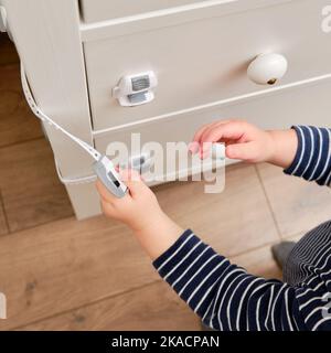 Das Baby öffnet das Kinderschloss in der geschlossenen Schublade des Schranks. Kleinkind Baby mit Hand öffnet Türschloss auf der Brust der Schubladen. Kind im Alter von einem Jahr und drei Stockfoto